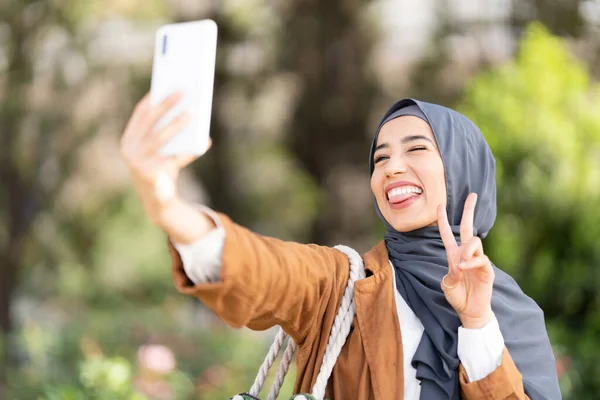 Muslimská Žena Šklebí Šťastný Zatímco Gestikuluje Vítězství Prsty Brát Selfie — Stock fotografie