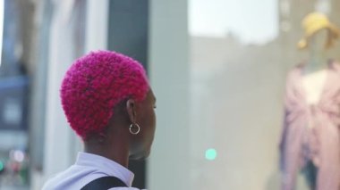 Afro woman with short pink hair looking in a shop window from the city street