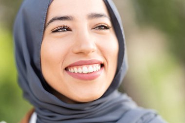 Açık havada rahat bir ifadeyle gülümseyen çekici bir müslüman kadının yakın plan portresi.