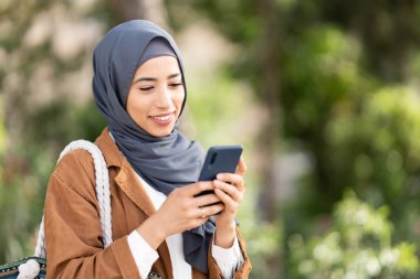Muslim woman using the mobile while smiling and standing in a park