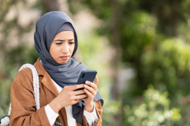 Muslim woman in hijab using the mobile phone with a sad expression of disgust in a park