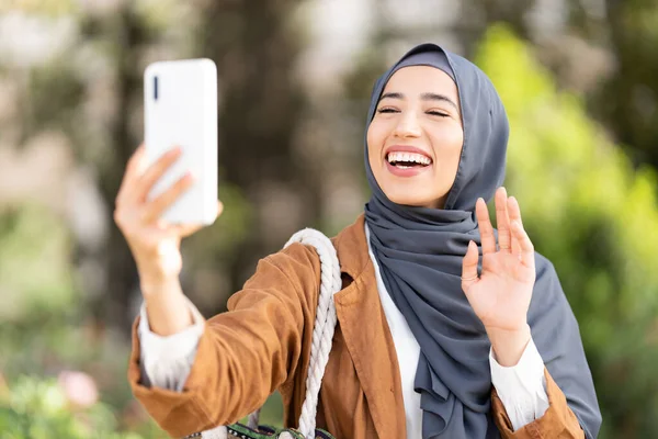 公園でビデオ通話をしながら 携帯電話のカメラに手を振ってヒジャーブのイスラム教徒の女性 — ストック写真