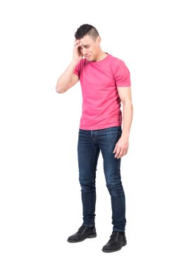 Pembe tişörtlü, kot pantolonlu, üzgün genç bir erkek. Alnına dokunuyor ve beyaz arka planda ağlarken hayal kırıklığıyla aşağıya bakıyor.