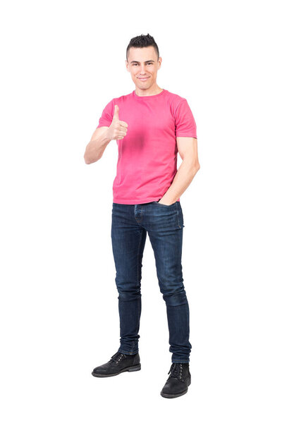 Full body of satisfied young male model with dark hair smiling and showing thumb up while standing against white background with hand in pocket and looking at camera