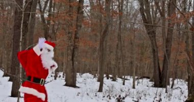 Kış ormanlarında yürürken, Noel Baba Noel hediyelerini taşıyarak elini sallıyor.