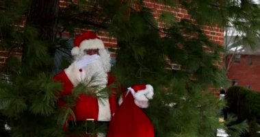 Noel Baba, Noel ağacının yanında duruyor. Elinde kırmızı bir çantayla çocuklara Noel için hediyeler var. Kar etrafında.