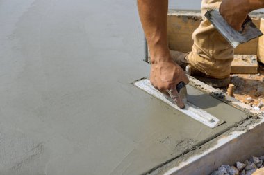 Bir inşaat işçisi, beton zemine alçı döşemeyi yumuşatırken elinde çelik bir mala tutuyor.