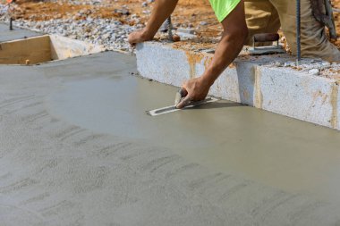 Çelik bir mala tutan bir inşaat işçisi var. Çelik bir malayla beton zemine beton yapıştırıyor.