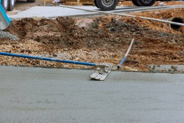 This construction worker is holding a steel trowel and smoothing out the plastering of new sidewalk on freshly laid concrete slab