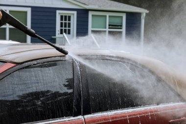 Adam arabasını yüksek basınçlı jetin altında yıkıyor. Arabayı temizlemek amacıyla su püskürtüyor.