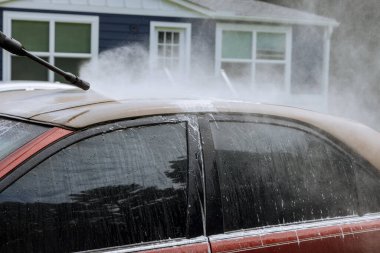 Adam yüksek basınçlı bir jetin altında araba yıkıyor. Temizlik maddesiyle su püskürtüyor.