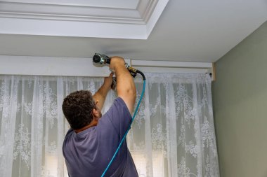 Yeni inşa edilen evde, tamirci, perdenin üstündeki ahşap kornişlere çivi çakıyor..
