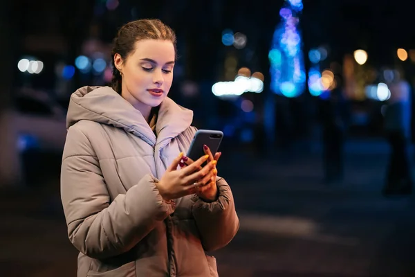 Woman ordering taxi using phone social media outside night evening street city lights winter cold weather. Christmas tree New Year atmosphere illumination copy space