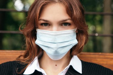 Dışardaki bankta oturan kadın bulutlu hava parkında maske takıyor. Bayan kendini damlayan su birikintilerinden korunmuş hissediyor. Boşluğu kopyala