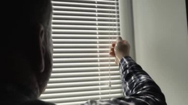 Erkek eli açık, bükme kolu açık, pencereye panjur kapatıyor, odasına bakıyor, güneş ışığı güneş evin dairesine giriyor..