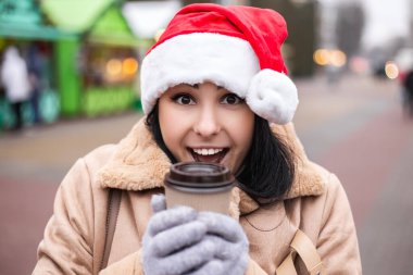 Genç, güzel kadın, kutlama eldivenleri sıcak içecek, kahve ya da çay, çevre dostu fincan. Soğuk hava, kış tatilleri fuarı, tatil beldesi. Yılbaşı şapkası Noel ağacı süslemeleri
