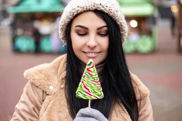 若いかなり笑顔を開いて口の白い歯の女性を祝うホールドロリポップ甘いキャンディーを食べています 寒い天候 冬の市場の休日の公正 旅行精神リゾート 新年のクリスマス飾り — ストック写真