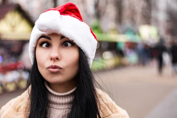 若いかわいい女性は 暖かいコートのミトンを祝うことをジェスチャー顔の顔をグリームを作る 寒い天候 冬の市場の休日の公正 旅行精神リゾート 新年の帽子クリスマスの装飾 — ストック写真