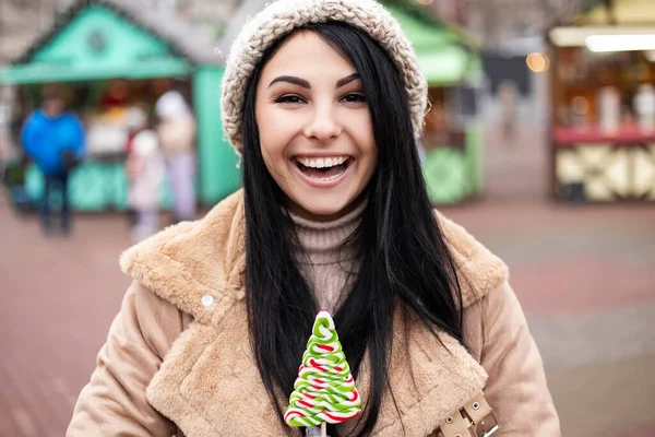 若いかなり笑顔を開いて口の白い歯の女性を祝うホールドロリポップ甘いキャンディーを食べています 寒い天候 冬の市場の休日の公正 旅行精神リゾート 新年のクリスマス飾り — ストック写真