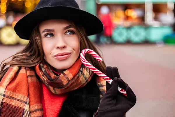 若いかなり笑顔の女性は暖かい羊の皮のコートを着て祝うロリポップ甘いストライプキャンディーを食べ保持しています 寒い天候 冬の市場の休日の公正 旅行リゾート 新年のクリスマス飾り — ストック写真
