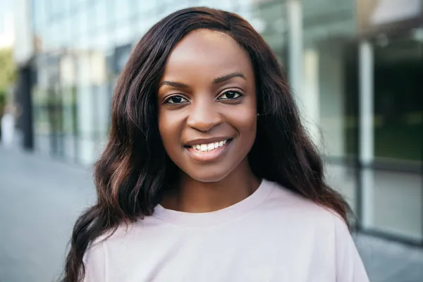 Kapalı portre güzel Afro-Amerikan siyah saçlı güzel kadın gülümseyen beyaz dişler takıyor sokak dışında kameraya bakıyor yaz tişörtü giymiş bulanık arka plan