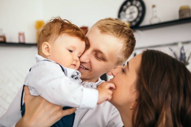 Küçük tatlı çocuk kızıl bebek anne babasıyla oynaşıyor ve oğlunu öpüyor. Yeni yürümeye başlayan çocuk evde tulum ve beyaz gömlek giyerek eğleniyor. Mutlu çocukluk, aile aşkı kavramı