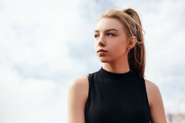 Woman standing outside dressed in sportswear preparing for training or running. Lady looking aside, sky with clouds on background.