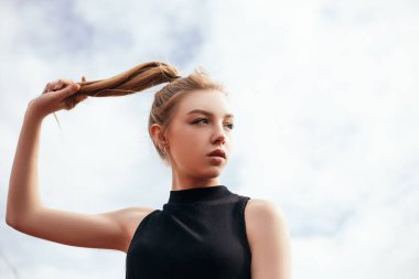 Woman standing outside dressed in sportswear preparing for training or running, keeps her pigtail. Lady looking aside, sky with clouds on background.