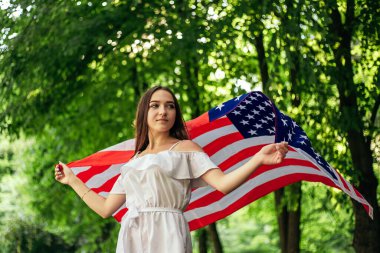 4 Temmuz kutlu olsun! Bağımsızlık Günü kutlaması. Vatansever kadın Amerikan bayrağına sarılmış rüzgarda sallanıyor ve yolda yürüyor. Yıldızlar ve çizgiler. Özgürlük kavramı.