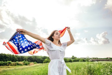 4 Temmuz kutlu olsun! Bağımsızlık Günü kutlaması. Vatansever kadın Amerikan bayrağına sarılmış rüzgarda sallanıyor ve sahada yürüyor. Yıldızlar ve çizgiler. Özgürlük kavramı.