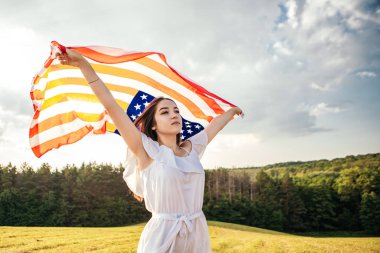 4 Temmuz kutlu olsun! Bağımsızlık Günü kutlaması. Vatansever kadın Amerikan bayrağına sarılmış rüzgarda sallanıyor ve sahada yürüyor. Yıldızlar ve çizgiler. Özgürlük kavramı.
