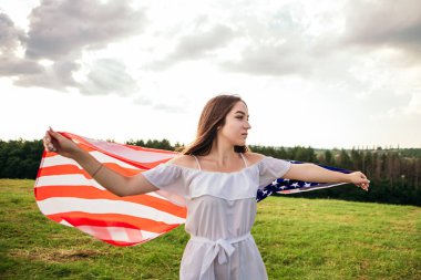 4 Temmuz kutlu olsun! Bağımsızlık Günü kutlaması. Vatansever kadın Amerikan bayrağına sarılmış rüzgarda sallanıyor ve sahada yürüyor. Yıldızlar ve çizgiler. Özgürlük kavramı.