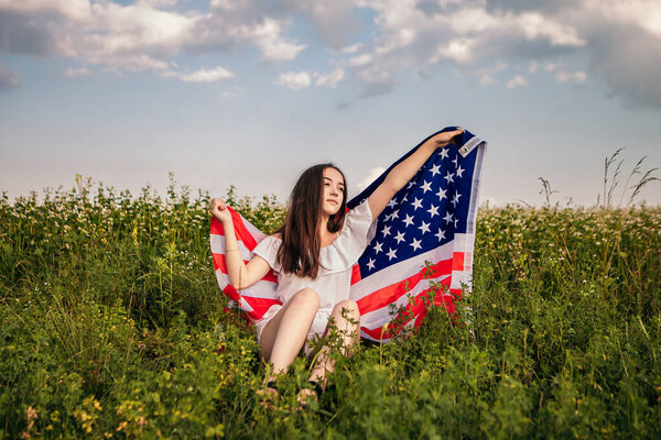 Happy 4th of July! Independence Day celebrating. Patriotic woman hold wrapped in american national flag waving on wind and walking on the field. Stars and stripes. Freedom concept.