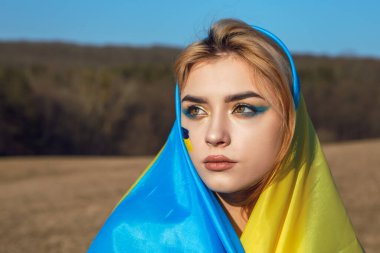 Ukrayna sarısı mavi bayrağa sarılı kadın portresi. Ukrayna 'nın ulusal sembolü yanakta. Makyajlı vatansever kadın sahada yürüyor. Ukrayna, destek ve dayanışma