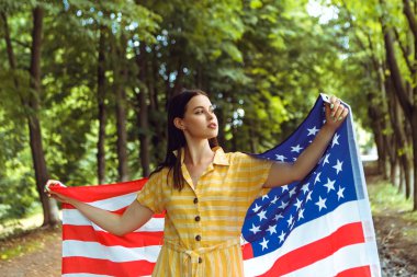 4 Temmuz kutlu olsun! Bağımsızlık Günü kutlaması. Vatansever kadın Amerikan bayrağına sarılmış rüzgarda sallanıyor ve yolda yürüyor. Yıldızlar ve çizgiler. Özgürlük kavramı.