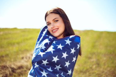 4 Temmuz kutlu olsun! Bağımsızlık Günü kutlaması. Vatansever kadın Amerikan bayrağına sarılmış rüzgarda sallanıyor ve sahada yürüyor. Yıldızlar ve çizgiler. Özgürlük kavramı.