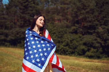 4 Temmuz kutlu olsun! Bağımsızlık Günü kutlaması. Vatansever kadın Amerikan bayrağına sarılmış rüzgarda sallanıyor ve sahada yürüyor. Yıldızlar ve çizgiler. Özgürlük kavramı.