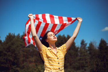 4 Temmuz kutlu olsun! Bağımsızlık Günü kutlaması. Vatansever kadın Amerikan bayrağına sarılmış rüzgarda sallanıyor ve sahada yürüyor. Yıldızlar ve çizgiler. Özgürlük kavramı.