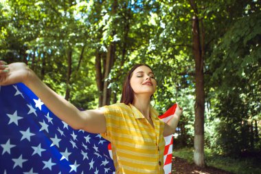 4 Temmuz kutlu olsun! Bağımsızlık Günü kutlaması. Vatansever kadın Amerikan bayrağına sarılmış rüzgarda sallanıyor ve yolda yürüyor. Yıldızlar ve çizgiler. Özgürlük kavramı.