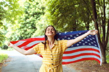 4 Temmuz kutlu olsun! Bağımsızlık Günü kutlaması. Vatansever kadın Amerikan bayrağına sarılmış rüzgarda sallanıyor ve yolda yürüyor. Yıldızlar ve çizgiler. Özgürlük kavramı.
