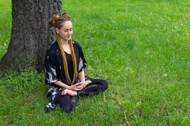 Rastalı yoga yapan kadın Siddhasana Ardha Padmasana Lotus 'un yeşil çimlerde poz çalışmasını yapıyor. Spor hocası egzersiz yapıyor, meditasyon yapıyor, gözleri kapalı.