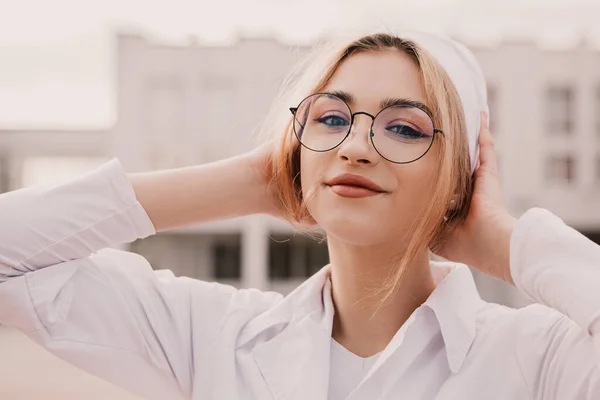 Hastane revirinin önünde duran genç bayan hemşire. Güzel doktor kadın beyaz bir elbise ve şapka giyip gözlük takıyor poz veriyor, gülümsüyor ve kameraya bakıyor..