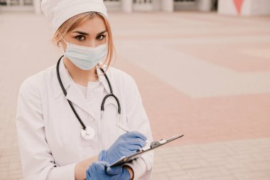 Revirin dışında duran genç bayan hemşire günlüğe hasta hastalık geçmişini yazıyor. Beyaz elbiseli doktor kadın, lastik mavi eldiven, steteskop ve maske doldurma kağıdı.