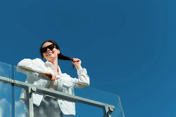 Portrait of smiling caucasian woman with white teeth standing near modern glass balcony railing. Perfect healthy smile with veneer. Blue clean sky background with copy space.