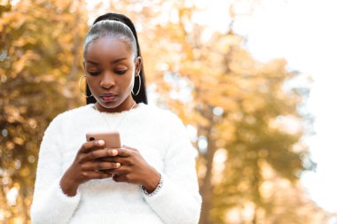 African pretty woman hold hand phone surfing internet chatting friends colleague send message standing outside autumn city park blur bokeh yellow leaves background