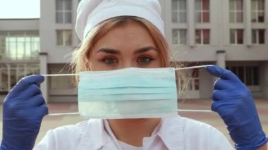 Young female nurse standing outside hospital infirmary. Gorgeous doctor woman dressed white medical gown and cap wear face disposable blue mask, looking at the camera.
