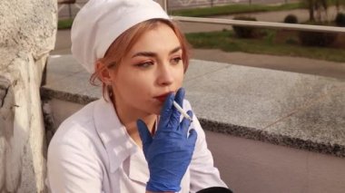 Dirty tired exhausted nurse ash on face sitting hospital infirmary smoking cigarette after hard working day or surgery. Doctor woman dressed white medical gown, rubber blue gloves have a rest due to stress