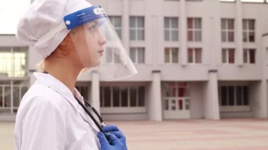 Hastane revirinin önünde duran genç bayan hemşire. Güzel doktor kadın beyaz bir elbise ve yüz koruyucu giymişti. Steteskop boynundaydı ve kameraya bakıyordu.