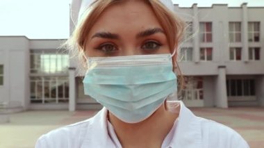 Young female nurse standing outside hospital infirmary. Gorgeous doctor woman dressed white medical gown and cap wear face disposable blue mask, looking at the camera.