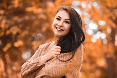 Close up portrait of a smiling happy toothy woman standing in autumn city park forest, looking at the camera open mouth with white teeth. Fall time, red orange and yellow leaves. Blur background